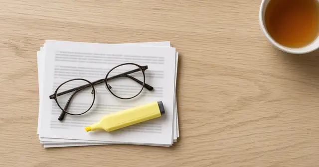 Seen from above: round reading glasses and a yellow highlighter on a stack of printed pages with unreadable text, next to a cup of tea on a light wooden table.
